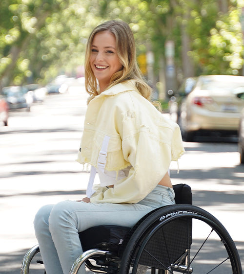 A picture of Catherine Elliott. She's sitting on a wheelchair smiling towards the camera. She is wearing vibrant yellow, spring clothing.
