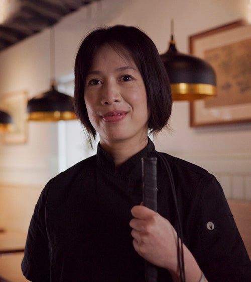 Chef Christine Ha holding a white cane. She is wearing a black chef apron and smiling at the camera.  